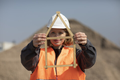 Frau mit reflektierender Weste und Schutzhelm, die einen Zollstock in Form eines Hauses hält, lizenzfreies Stockfoto