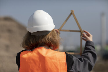 Rear view of woman wearing reflective vest and hard hat holding pocket rule in shape of a house - SGF02360