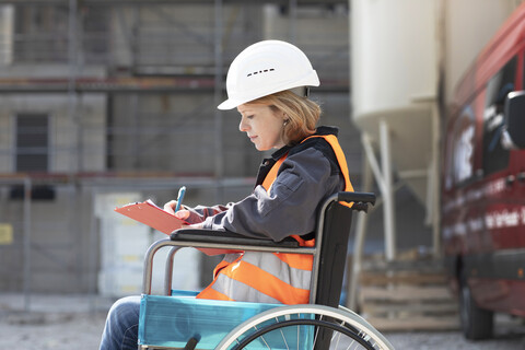 Woman wearing reflective vest and hard hat sitting in wheelchair taking notes stock photo