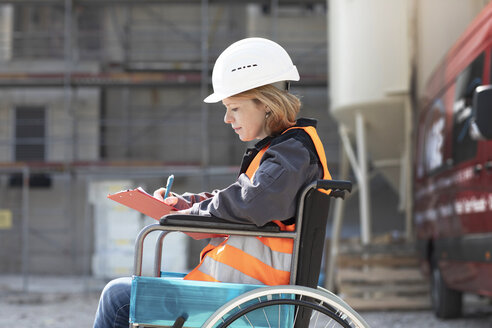 Frau mit Warnweste und Schutzhelm sitzt im Rollstuhl und macht Notizen - SGF02361
