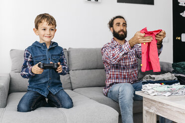 Portrait of smiling little boy playing computer game while his father folding laundry - JRFF03008