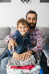 Portrait of father and son sitting together on the couch folding laundry - JRFF03010