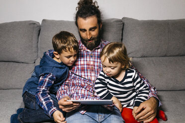 Father sitting on the couch with his children watching movies on digital tablet - JRFF03014