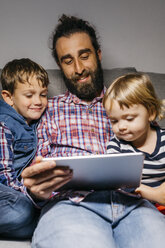 Portrait of father sitting on the couch with his children watching movies on digital tablet - JRFF03015