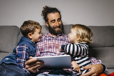 Happy father sitting on the couch with his children using digital tablet - JRFF03016