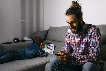 Vater sitzt auf der Couch und schaut auf sein Smartphone, während sein Sohn im Hintergrund ein digitales Tablet benutzt - JRFF03017