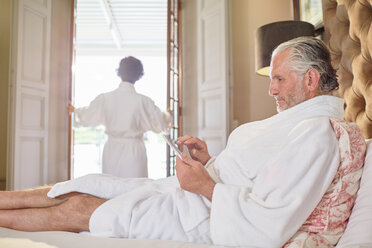 Älterer Mann im Bademantel mit digitalem Tablet auf einem Hotelbett - CAIF23150