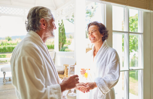 Glückliches reifes Paar in Bademänteln trinkt Mimosen an der Balkontür eines Hotels - CAIF23152