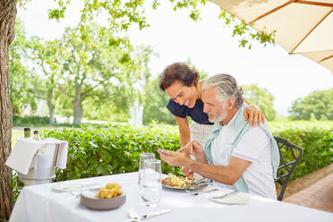 Älteres Paar beim Essen, das am Terrassentisch sein Smartphone benutzt - CAIF23172