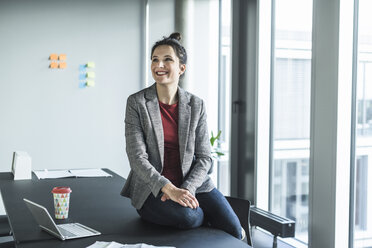 Smiling businesswoman sitting on desk in office - UUF17083