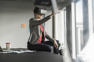 Businesswoman sitting on desk in office wearing VR glasses - UUF17090