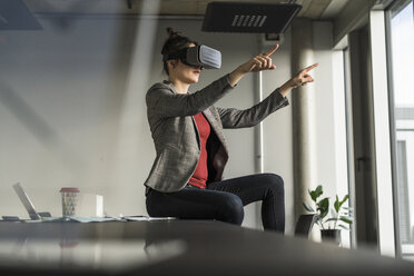 Businesswoman sitting on desk in office wearing VR glasses - UUF17091