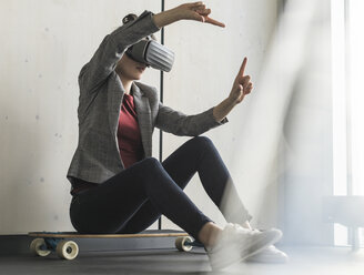 Geschäftsfrau sitzt auf einem Skateboard im Büro und trägt eine VR-Brille - UUF17092