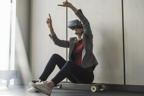 Geschäftsfrau sitzt auf einem Skateboard im Büro und trägt eine VR-Brille, lizenzfreies Stockfoto