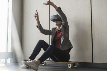 Geschäftsfrau sitzt auf einem Skateboard im Büro und trägt eine VR-Brille - UUF17093