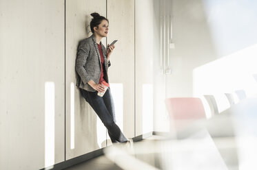 Geschäftsfrau lehnt an einer Wand im Büro und benutzt ein Mobiltelefon - UUF17094