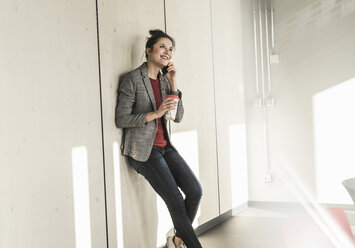 Lächelnde Geschäftsfrau lehnt an einer Wand im Büro und spricht mit einem Handy - UUF17096