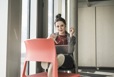 Businesswoman sitting on chair in office with laptop and earphones - UUF17103