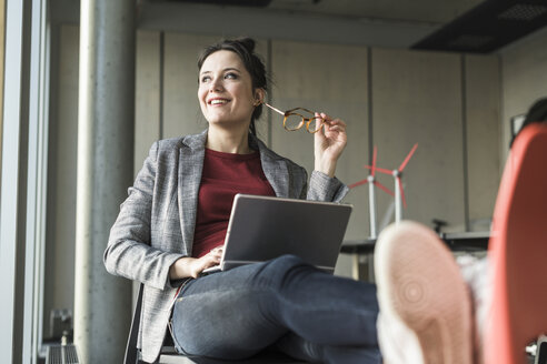 Lächelnde Geschäftsfrau auf einem Stuhl im Büro sitzend mit Laptop und Blick zur Seite - UUF17104