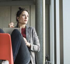 Businesswoman sitting on chair in office with laptop looking sidways - UUF17105