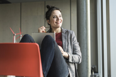 Smiling businesswoman sitting on chair in office with laptop looking sidways - UUF17106