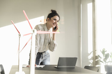 Geschäftsfrau mit Windkraftanlagenmodellen und Laptop auf dem Schreibtisch im Büro, die mit dem Handy telefoniert - UUF17110