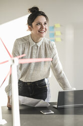 Smiling businesswoman with wind turbine model and laptop on desk in office - UUF17111