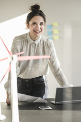 Portrait of smiling businesswoman with wind turbine model on desk in office - UUF17112