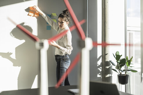 Geschäftsfrau im Büro, die Haftnotizen an die Wand klebt, mit Windradmodellen auf dem Schreibtisch - UUF17116