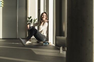 Lächelnde Geschäftsfrau sitzt auf dem Skateboard am Fenster im Büro - UUF17127