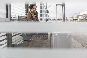 Lächelnde Geschäftsfrau mit Gepäck sitzt am Bahnhof und schaut auf ihr Handy - UUF17137