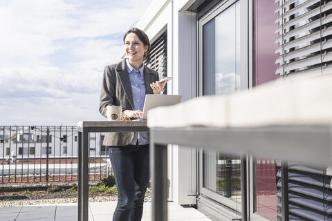 Smiling businesswoman with cell phone and laptop on roof terrace stock photo