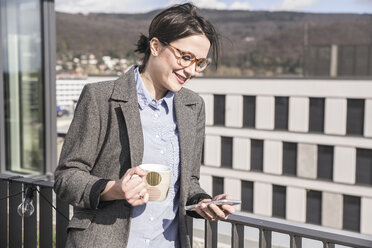 Smiling businesswoman with cell phone and cup of coffee on roof terrace - UUF17145