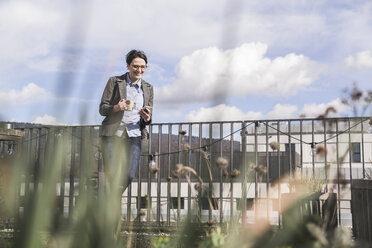 Smiling businesswoman with cell phone and cup of coffee on roof terrace - UUF17147