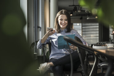 Smiling businesswoman sitting in office with paper and cell phone - UUF17151