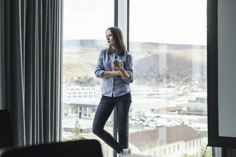 Geschäftsfrau mit Handy steht am Fenster im Büro, lizenzfreies Stockfoto