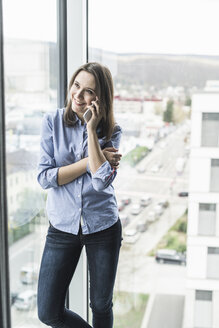 Lächelnde Geschäftsfrau mit Handy am Fenster im Büro stehend - UUF17159