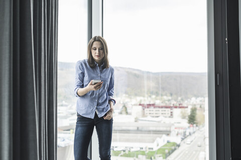 Geschäftsfrau mit Handy steht am Fenster im Büro, lizenzfreies Stockfoto