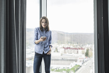 Businesswoman with cell phone standing at the window in office - UUF17160