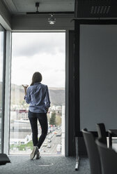 Rückansicht einer Geschäftsfrau mit Handy, die am Fenster eines Büros steht - UUF17161