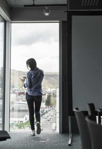 Rückansicht einer Geschäftsfrau mit Handy, die am Fenster eines Büros steht, lizenzfreies Stockfoto