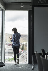 Rückansicht einer Geschäftsfrau mit Handy, die am Fenster eines Büros steht - UUF17162