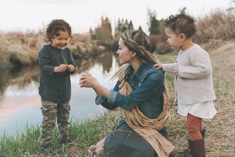Mutter und Kinder spielen mit einem Grashalm an einem Fluss, lizenzfreies Stockfoto