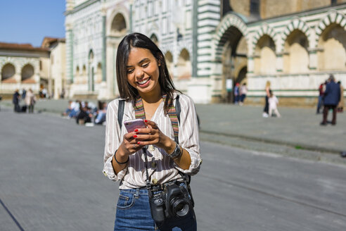 Italien, Florenz, Porträt eines glücklichen jungen Touristen mit Kamera und Rucksack, der auf sein Handy schaut - MGIF00338