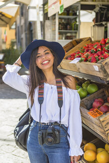 Italien, Florenz, Porträt eines glücklichen jungen Touristen mit Kamera vor einem Lebensmittelgeschäft - MGIF00359