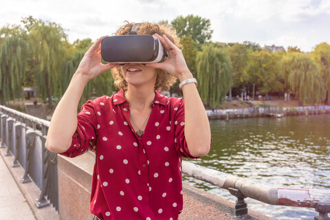Junge Frau mit VR-Headset auf Brücke, Fluss im Hintergrund, Berlin, Deutschland, lizenzfreies Stockfoto