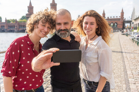 Mann und Freundinnen machen Selfie mit Smartphone auf Brücke, Fluss und Gebäude im Hintergrund, Berlin, Deutschland, lizenzfreies Stockfoto