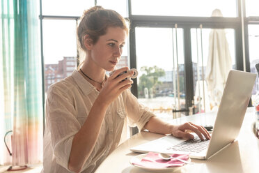 Young woman using laptop and having coffee in office on sunny day - CUF49967