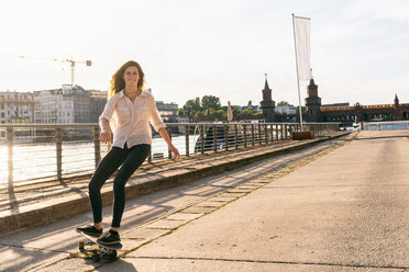 Young woman skateboarding on bridge, river and buildings in background, Berlin, Germany - CUF49970