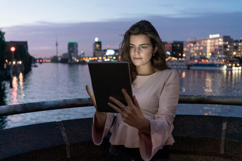Junge Frau mit digitalem Tablet auf einer Brücke, Fluss und Stadt im Hintergrund, Berlin, Deutschland, lizenzfreies Stockfoto